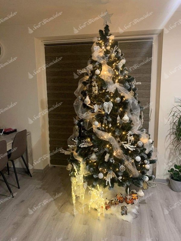 Umleý vianočný stromček so zasneženými koncami vetvičiek , ozdobený bielymi ozdobami a vianočnou stuhou s bielym koberčekom pod stromček.